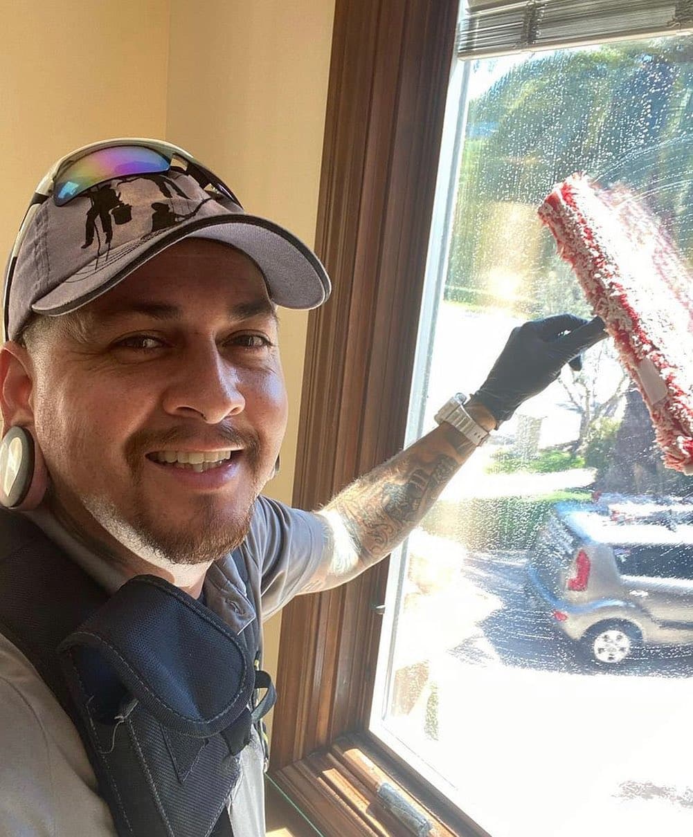 A man in a baseball cap and gloves holding a window cleaner.